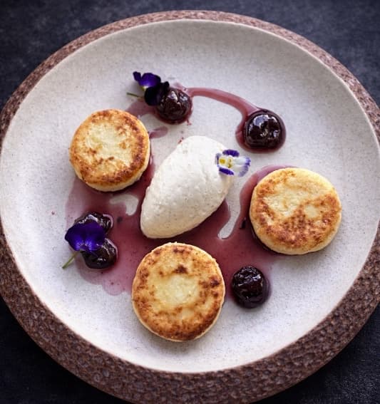 Syrniki  — Soft cottage cheese pancakes served with mascarpone-banana cream and cherry compote. Delicate, cream